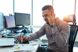 An actuary sits in front of a computer screen with print outs of graphs. He is smiling and talking on the phone.