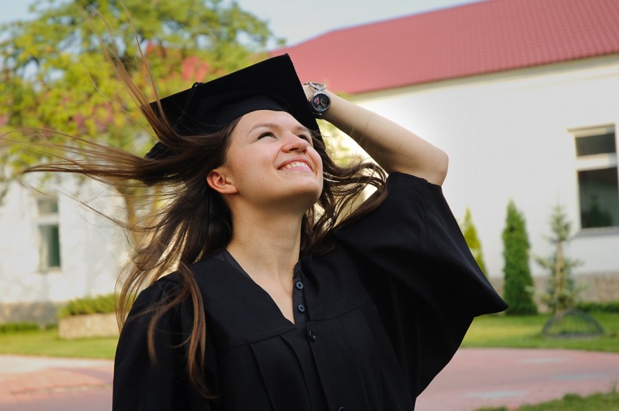 A young woman wearing a graduation cap and gown looks to the sky as wind blows her hair